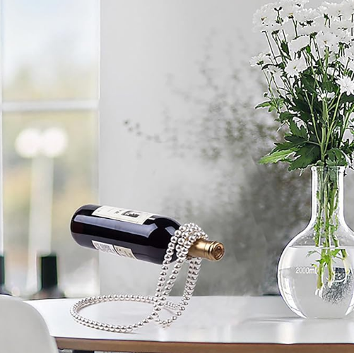 Porte-bouteille flottant en perles blanches, soutenant une bouteille de vin rouge sur une table blanche design, à côté d’un vase transparent rempli de fleurs blanches. Mise en scène élégante dans un intérieur lumineux et minimaliste.
